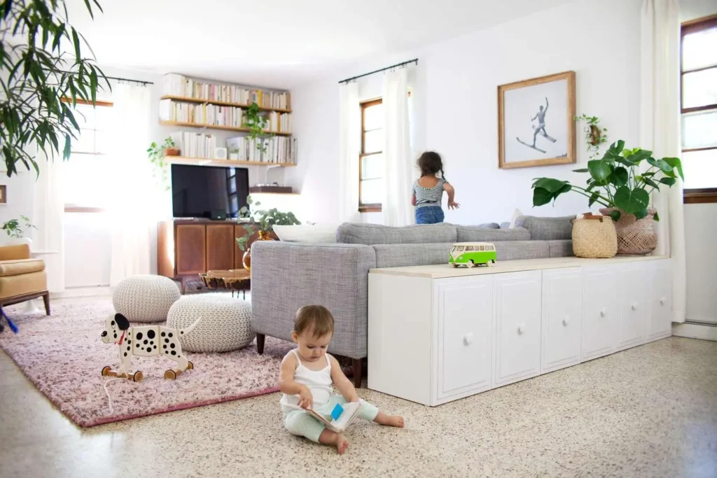 Bright living room with two toddlers playing: one on the floor reading a book, the other near the sofa; a Dalmatian toy on a pink rug, plants, and shelves.