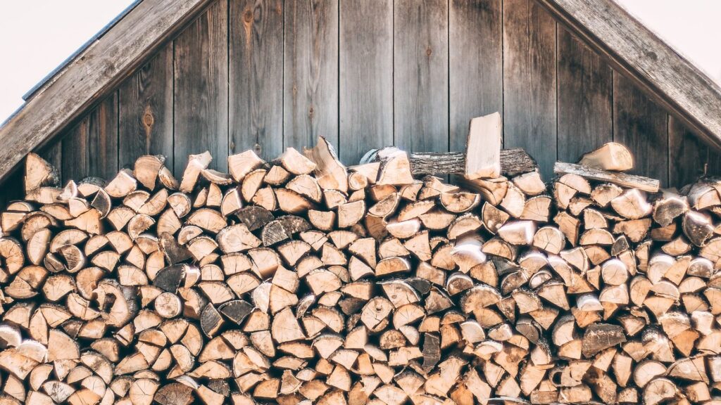 Polttopuut pinottuina tuulettuvaan puuvajaan - TaloEkspertti Stack of chopped firewood neatly stacked against a weathered wooden shed wall under a peaked roof.